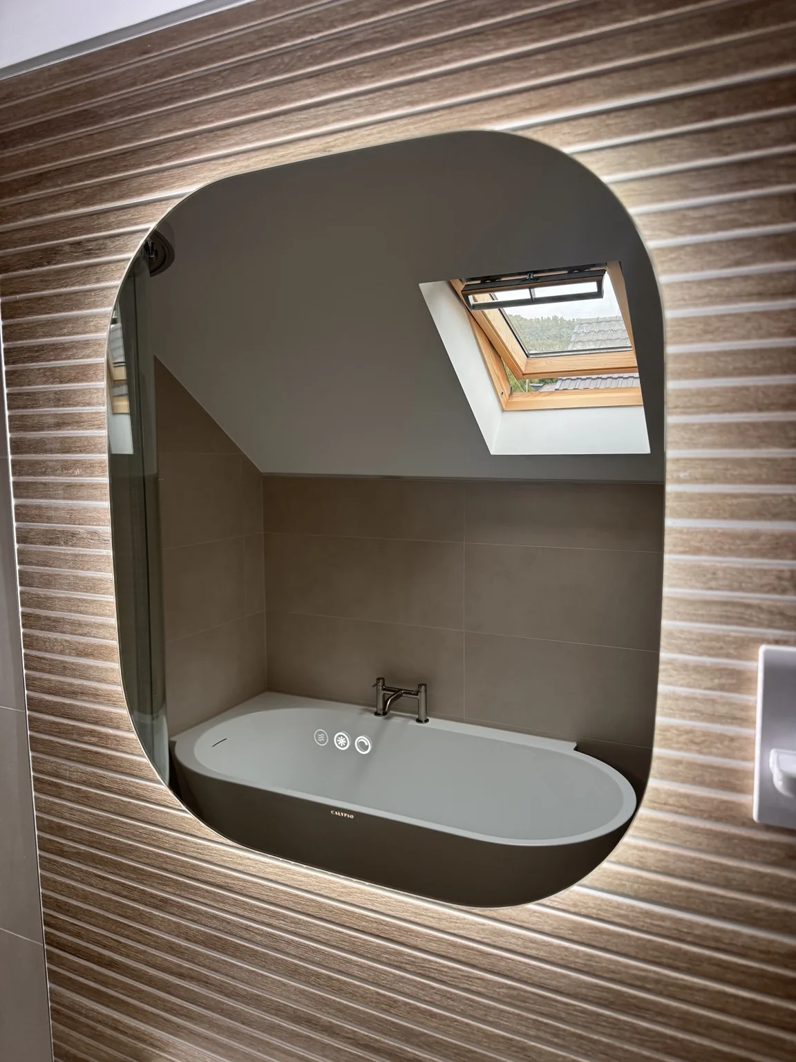 Illuminated mirror reflecting the semi-freestanding bath beneath the roof window